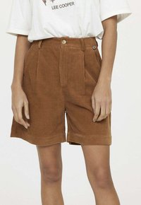 Shorts en velours côtelé marron avec une coupe ajustée, présentant des plis, un ourlet coutu et une fermeture à bouton. Portés avec un t-shirt graphique.