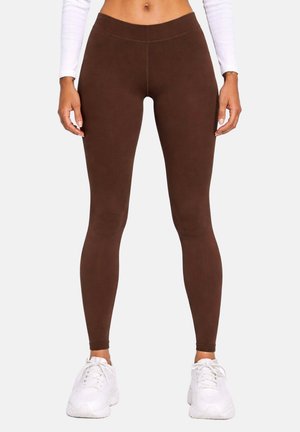 Femme portant des leggings marron ajustés et des baskets blanches, debout devant un fond uni avec les mains détendues le long du corps.
