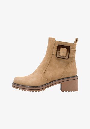 Stivaletto in suede beige con dettaglio a fibbia quadrata, tacco spesso e strutturato, e punta arrotondata. Presenta un grosso anello sul retro per facilitare l'indossamento.