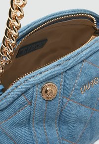 Denim-Handtasche mit goldenem Kettenriemen, gestepptem Textil und orangefarbenen Nähten. Verfügt über einen markierten Metallknopf und ein beiges Innenfutter.