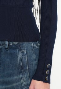 Tmavomodrý rebrovaný sveter s dlhými rukávmi, zdobený zlatými gombíkmi na manžetách, v kombinácii s vyblednutými džínsami.