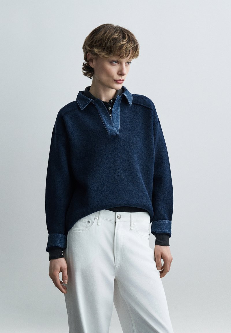 Jeune personne aux cheveux courts portant un pull bleu marine avec col et poignets en denim, associé à un pantalon blanc taille haute, debout devant un fond gris.