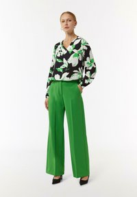 Bluse mit Blumenmuster in Schwarz, Weiß und Grün mit V-Ausschnitt; weite grüne Hose; schwarze High Heels; glattes Material, lockere Passform.