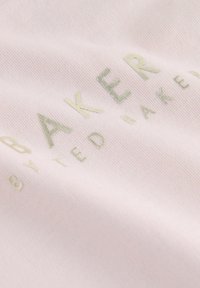 Lys pink stof med en blød tekstur, der har forhøjede, metalliske guldtekster, der står "BAKER by Ted Baker." Enkel og moderne design.