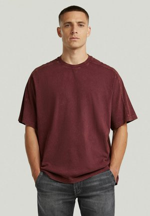 Uomo con capelli corti che indossa una t-shirt color bordeaux sbiadita a taglio morbido e jeans neri slavati, in piedi con le mani nelle tasche davanti a uno sfondo semplice.