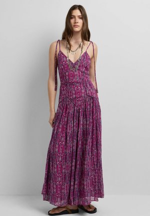 Femme portant une robe maxi sans manches, violette à motifs, avec bretelles spaghetti et un collier en argent à plusieurs couches sur un fond gris uni.