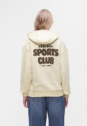 Човек със светла коса, носещ светложълта качулка с текст "ellesse SPORTS CLUB since 1959" на гърба и сини дънки.