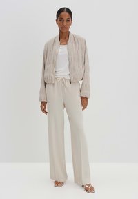 Hellbeige Bomberjacke mit strukturierten horizontalen Streifen, getragen über einem weißen T-Shirt und passenden beigen weit geschnittenen Hosen. Silberne Sandalen runden den Look ab.