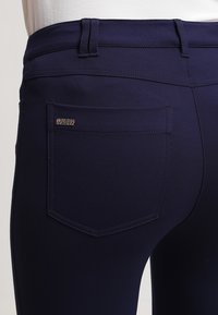 Marinblå tights med en slät textur, har bakfickor och en märkesetikett på den högra fickan. Sytt detaljarbete längs midjebandet.