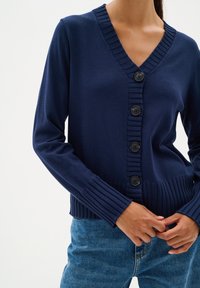 Marinblå stickad cardigan med V-ringning, fem svarta knappar och ribbade ärmslut och nederkant. Har en slät textur och figurnära design.
