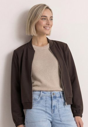 Ung kvinde med kort blondt hår har en brun lynlåstrøje på over en beige sweater og lyseblå højttaljede jeans, smiler let.