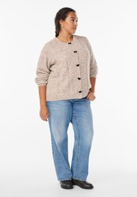 Jasnobrązowy kardigan o teksturze z czarnymi guzikami, zestawiony z jasnoniebieskimi spodniami o szerokich nogawkach i czarnymi botkami za kostkę, stojący na białym tle.