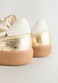 Goldfolien-Sneaker mit weißem, strukturiertem Stoff an der Ferse. Verfügt über ein glattes Finish, beige Sohle und abgesteppten Akzente für zusätzliche Details.