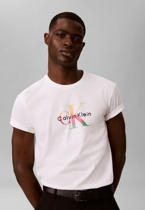 Mężczyzna ubrany w białą koszulkę Calvin Klein z wielokolorowym logo, czarne spodnie i czarny teksturowany pasek na jednolitym szarym tle.