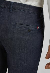 Donkerblauwe spijkerbroek met een knoop achterzak en een klein rood, wit en blauw stoffen label, gedragen met een wit topje.