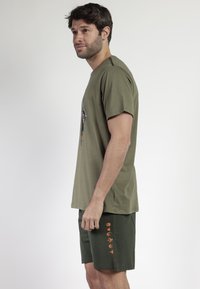 Hombre con camiseta gráfica verde oliva y pantalones cortos verdes oscuros con símbolos naranjas se presenta en perfil contra un fondo claro y liso.