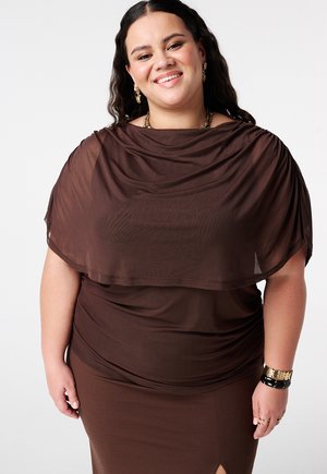 Femme souriante aux longs cheveux bruns portant un châle marron transparent sur une robe marron moulante, accessoirisée avec des bijoux et des bracelets en or.