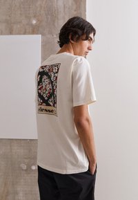 Cremefarbener Baumwoll-T-Shirt mit floraler Grafik auf der Rückseite in Rot, Gelb und Schwarz. Lockere Passform mit kurzen Ärmeln.