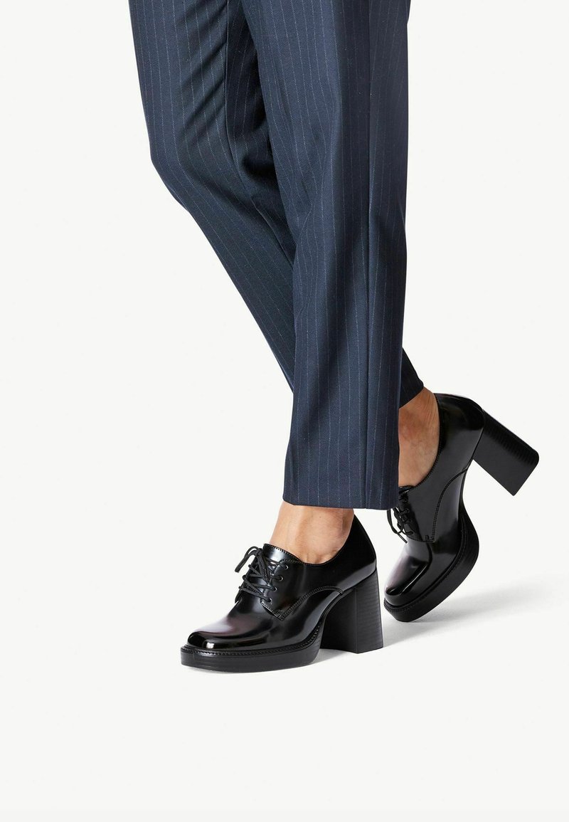 Personne portant un pantalon à fines rayures foncées et des chaussures noires brillantes à lacets avec talon bloc, debout jambes croisées sur fond blanc.