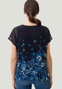 Blouse florale bleu marine à manches courtes, avec un motif de fleurs bleues varié et une texture fluide. Ourlet légèrement plus long à l'arrière.