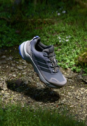Harmaa ja sininen Adidas Terrex -vaelluskenkä leijuu kivisen, nurmisen ulkopinnan yläpuolella auringonvalossa ja varjoissa.