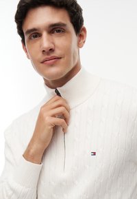 Mann mit kurzen dunklen Haaren, der einen weißen kabelgestrickten Pullover mit Reißverschluss trägt, hält den Reißverschluss nahe am Kragen vor einfarbigem Hintergrund.