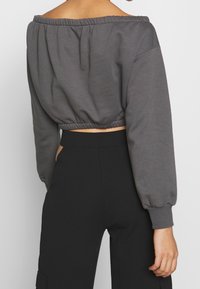 Sudadera gris corta con diseño de hombro al descubierto, dobladillo elástico y mangas largas abullonadas. Combinada con pantalones negros de tiro alto.