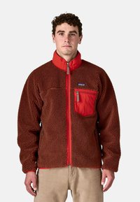 Uomo che indossa una giacca in pile con zip marrone e rossa, con tasca sul petto e pantaloni beige, in piedi contro uno sfondo semplice.