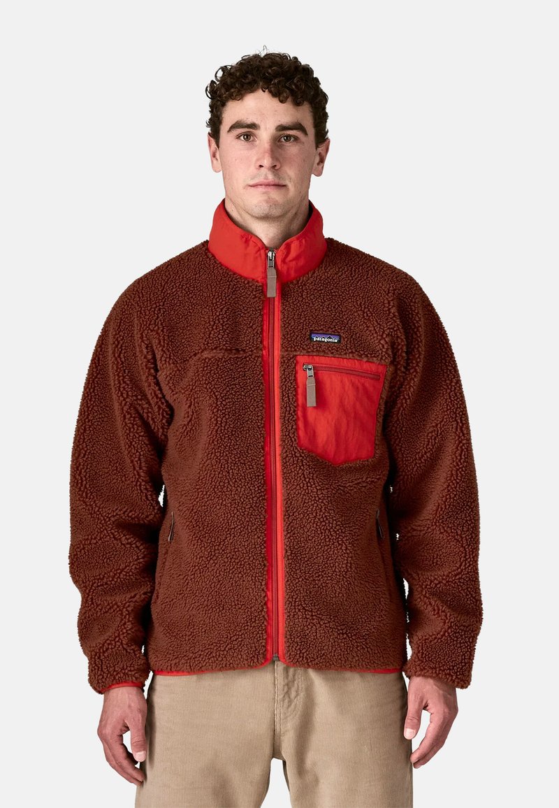 Uomo che indossa una giacca in pile con zip marrone e rossa, con tasca sul petto e pantaloni beige, in piedi contro uno sfondo semplice.