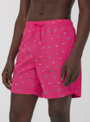 Uomo che indossa pantaloncini da bagno rosa acceso con motivi di squalo, pesci e onde blu, in piedi contro uno sfondo chiaro e uniforme.