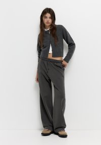 Cardigan gri din tricot peste un top scurt alb, asortat cu pantaloni lejeri gri din bumbac. Ținuta are o croială lejeră și un elastic la talie.