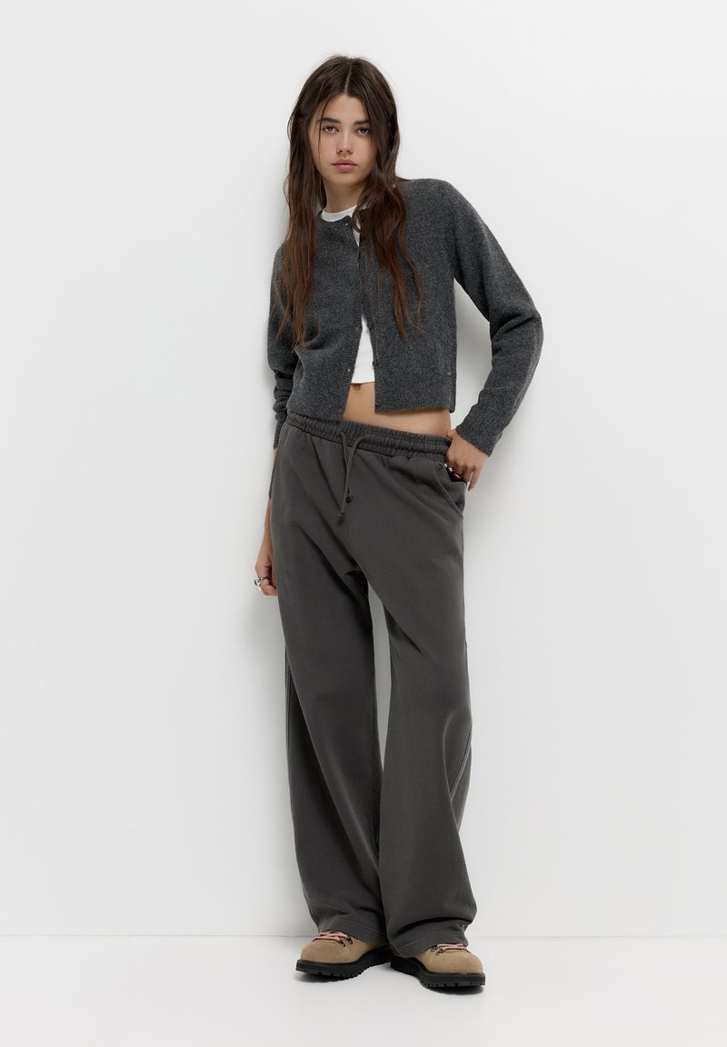 Cardigan gri din tricot peste un top scurt alb, asortat cu pantaloni lejeri gri din bumbac. Ținuta are o croială lejeră și un elastic la talie.