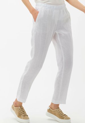 Person, die weiße Leinenhose mit elastischem Bund und beige Sneaker trägt, steht mit einer Hand in der Tasche vor weißem Hintergrund.