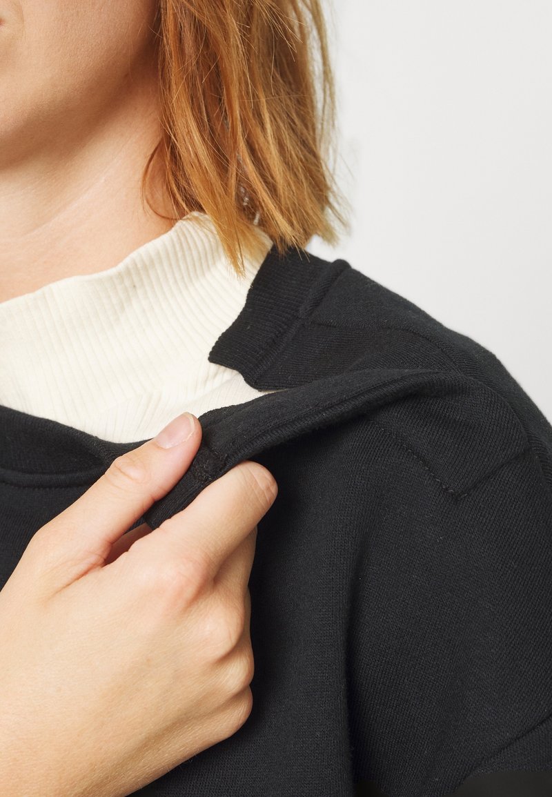 Une personne aux cheveux courts roux soulève un vêtement noir pour montrer un fermoir magnétique sur l'épaule, porté sur un t-shirt blanc côtelé.