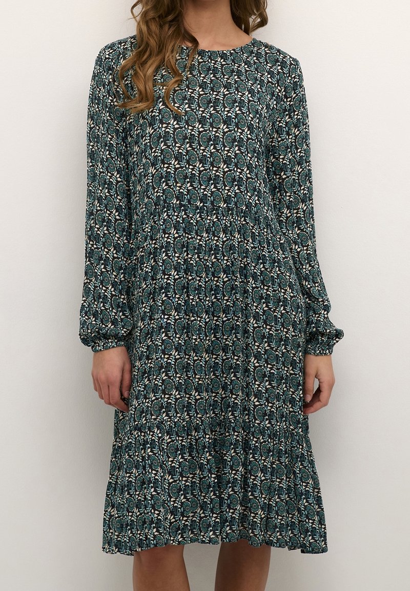 Femme portant une robe à manches longues, longueur genou, avec un motif floral vert foncé et une jupe froncée, debout devant un fond uni.