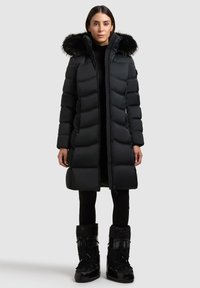 Svart dunjacka med luva kantad med päls, med ett quiltat mönster, dragkedja framtill och sidofickor. Matchas med svarta, texturerade stövlar.