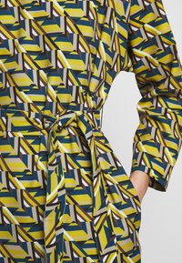 Vêtement à manches longues ceinturé avec motif géométrique jaune, bleu, marron et blanc, avec une main dans la poche latérale.