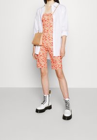 Combinaison orange à motifs floraux avec des shorts, assortie à une chemise blanche oversize et un sac à main en cuir beige embossé crocodile. Bottes de combat noires à lacets.