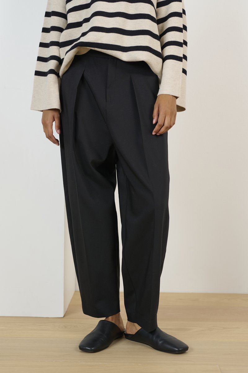 Pantalons plissés noirs, taille haute et coupe ample, associés à un pull surdimensionné à rayures beige et noires, et des chaussures noires à enfiler.