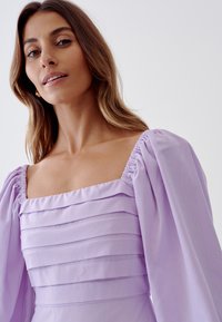 Lilac dress with a square neck, featuring tiered pleats on the bodice and puffed sleeves. Made of smooth fabric with a subtle texture.