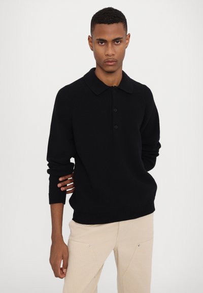 Polo en maille noire avec col, patte à trois boutons et poignets côtelés. Porté sur un pantalon de couleur claire. Design simple et texturé.