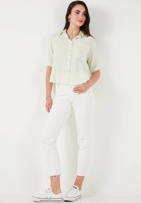 Camicia a maniche corte verde chiaro con bottoni e taschino sul petto, abbinata a jeans skinny bianchi e sneakers bianche con dettagli rossi.