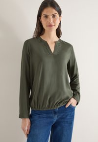 Blouse à manches longues vert olive avec un col en V et un ourlet froncé. Confectionnée dans un tissu lisse, avec une coupe décontractée et des manches retroussables.