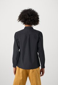 Black long-sleeve shirt with a buttoned collar, slight matte texture, and a central back pleat. Paired with yellow shorts.