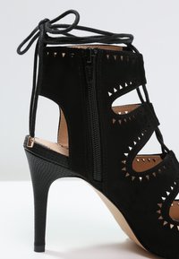 Sandale noire à talon haut avec fermeture éclair latérale, motifs triangulaires découpés, laçage à la cheville et talon texturé, présentée sur fond uni.