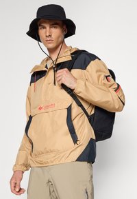 Beige og marineblå vindjakke med justerbar hette, frontlomme og Columbia-logo. Båret med en svart bøttehatt og ryggsekk.