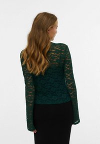Haut à manches longues en dentelle verte avec un motif floral, en tissu semi-transparent, coupe ajustée et encolure ronde, porté sur une jupe noire.