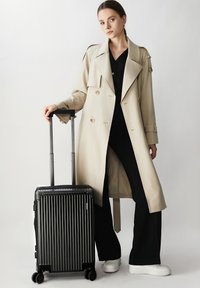 Abrigo trinchera beige con grandes solapas, combinado con pantalones negros y zapatillas blancas, al lado de una maleta negra con surcos y ruedas.