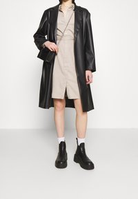 Mustat nahkatakki beige-napilla varustetun mekon päällä, yhdistettynä mustiin korkokenkisiin nilkkasaappaisiin ja pieneen mustaan käsilaukkuun, jossa on ketjuhihna.