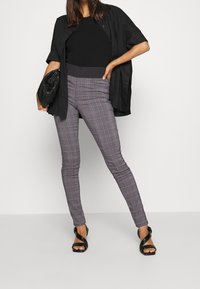 Pantalones de cuadros grises en forma de campana con sutiles acentos rojos, combinados con una blusa negra ajustada y un bolso de mano negro de gran tamaño, llevados con tacones negros de tiras.